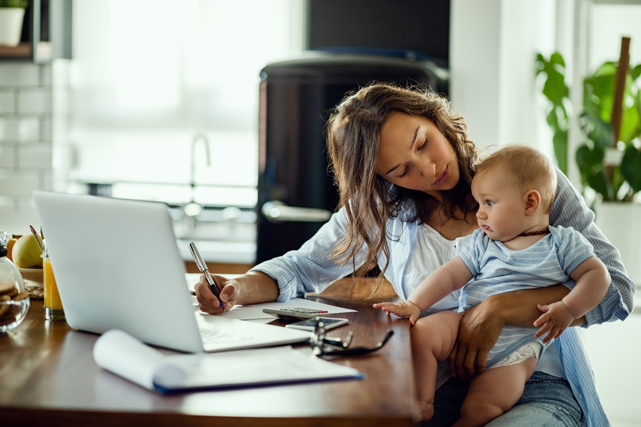 A mother holding her baby while writing beside a laptop at home
